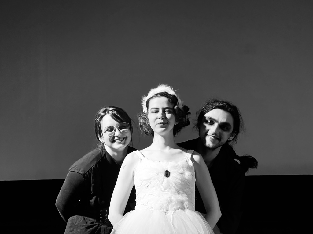 Image of three actors smiling at the camera. The actor in front is dressed as a ballerina, and two figures in black are looking over her shoulder.
